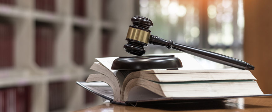 Judge's gavel resting on a law book on a desk, with blurred bookshelves and warm sunlight in the bac